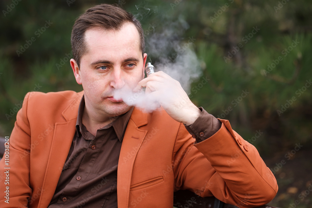 A young man sits on a bench and smokes an electronic cigarette. A young man sits on a bench and smokes an electronic cigarette.