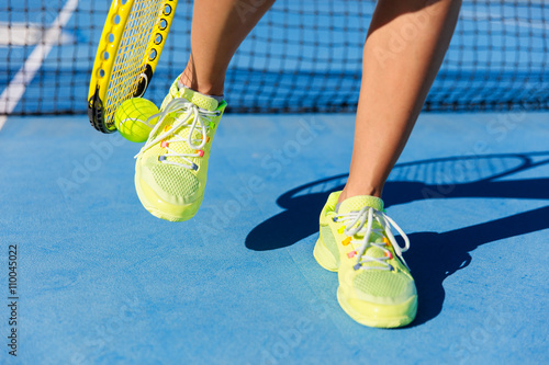 Sports athlete picking up ball with tennis racket. Female player using a technique with her running shoes to pick up during game on blue hard court. Closeup of feet, neon yellow fashion footwear. © Maridav