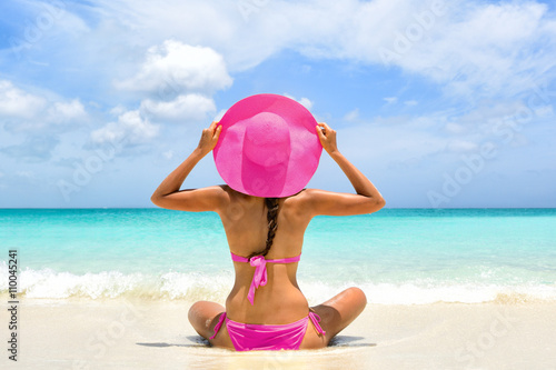 Cute pink bikini beachwear woman relaxing in perfect paradise destination on beach travel vacation. Girl from the back holding fashion straw floppy hat sitting on sand looking a turquoise ocean.