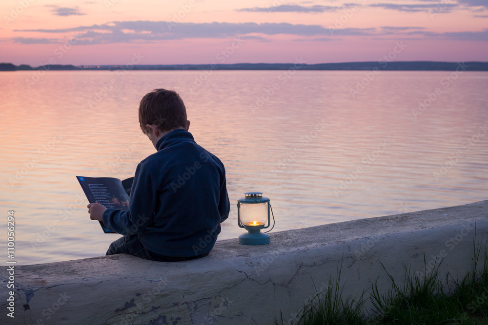 Fototapeta premium child reading a book with lantern at sunset near water