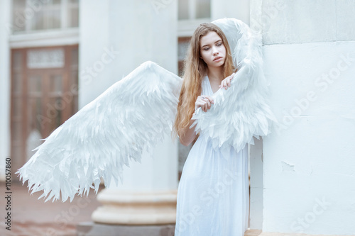 Young woman with white wings is standing near the column. The girl hid wing