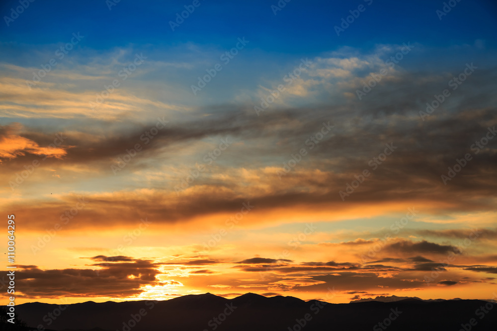 Sun behind dark mountain silhouettes, with colorful sky and clouds
