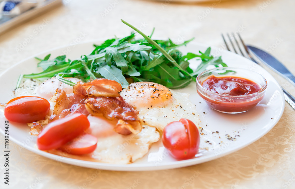Breakfast on plate - fried eggs, fried bacon, cherry tomatoes, ruccola, black ground pepper and red sauce