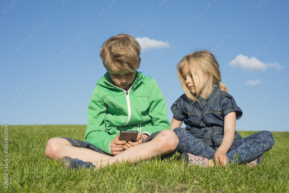 Happy blond children using smartphone (watching movie or playing game) sitting on the grass. Brother and sister. Summertime Happy blond children using smartphone (watching movie or playing game) sitting on the grass. Brother and sister. Summertime