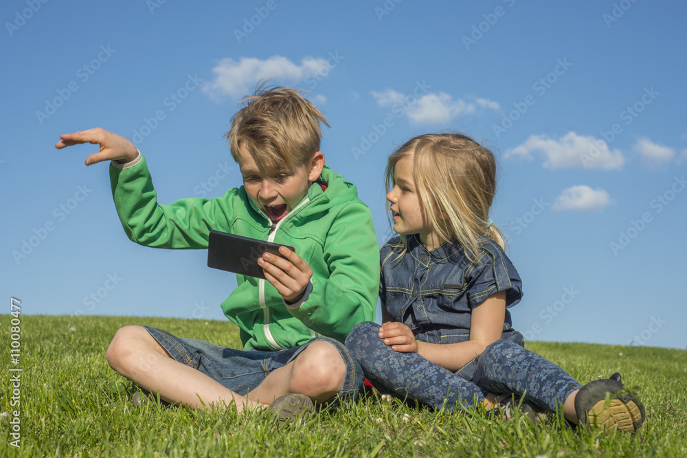Happy blond children using smartphone (watching movie or playing game) sitting on the grass. Brother and sister. Summertime Happy blond children using smartphone (watching movie or playing game) sitting on the grass. Brother and sister. Summertime