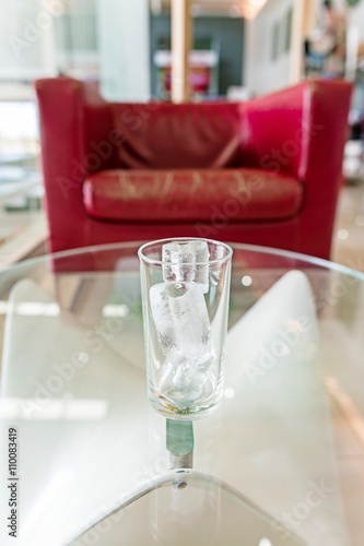 glass with ice cubes on a glass table and blurry red sofa background 