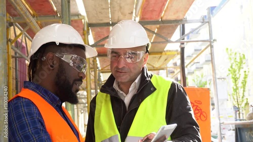Architect wearing yellow safety vest and goggles providing instructions to a colleague with orange vest and tablet on construction site