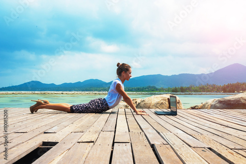 Woman in office suit doing yoga on the wooden floor with laptop. Young Lady has practice on the beach in front of sea view.