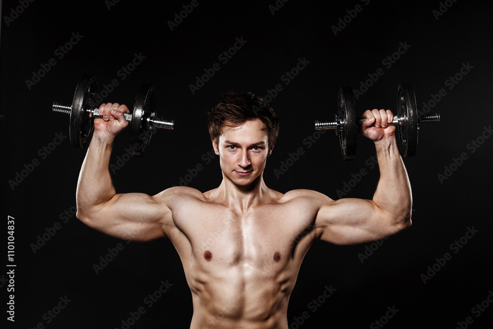 Handsome athletic man with dumbbells confidently looking forward. Strong bodybuilder with six pack, perfect abs, shoulders, biceps, triceps and chest. Great fitness body. Ideal for commercial 