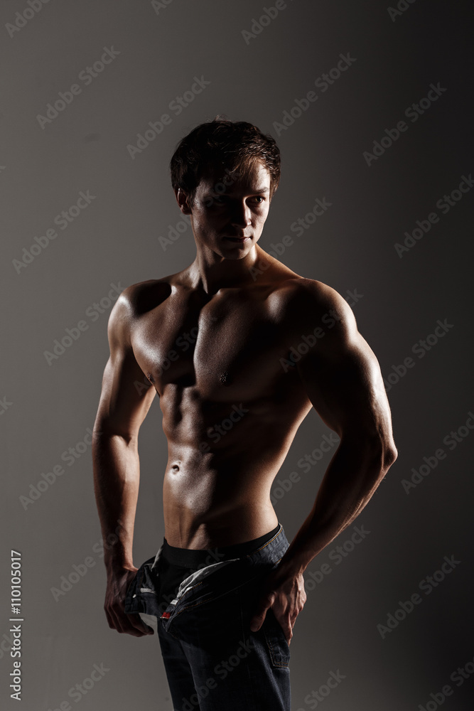 Handsome athletic man looking side in unbuttoned jeans. Strong bodybuilder with six pack, perfect abs, shoulders, biceps, triceps and chest. Great fitness body. Ideal for commercial. Gray background