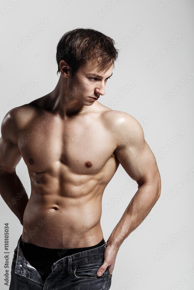 Handsome athletic man looking side in unbuttoned jeans. Strong bodybuilder with six pack, perfect abs, shoulders, biceps, triceps and chest. Great fitness body. Ideal for commercial. Gray background