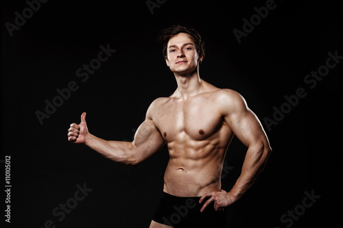 Muscular and fit young bodybuilder fitness male model showing thumb-up and posing over black background. Perfect fitness body. Ideal for commercial.