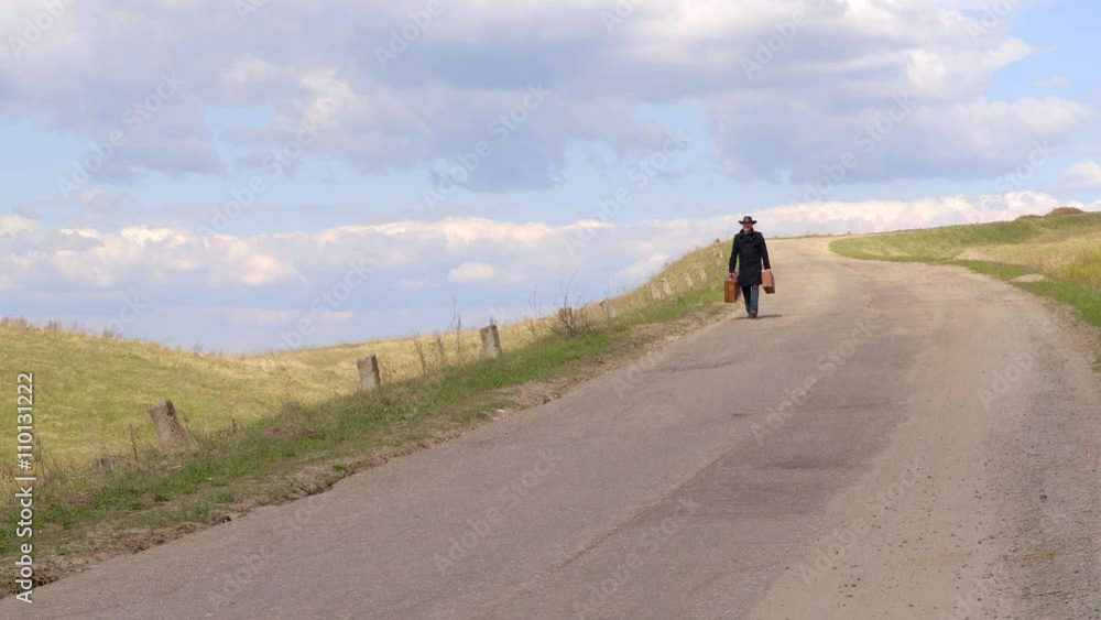 The man walking along the road with two old suitcases on the grass in the summer The man walking along the road with two old suitcases on the grass in the summer