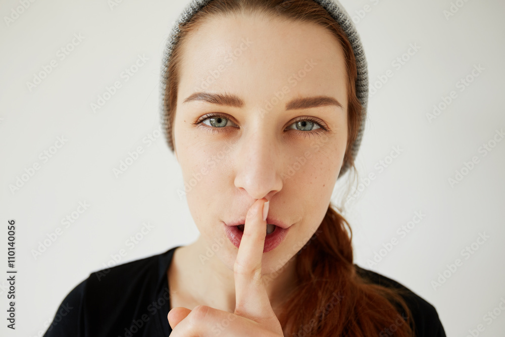 Headshot of young redhead woman with finger at her lips showing hush sign asking to be quiet. Young mother asking to keep silence while her little children asleep. Human face expressions and emotions Headshot of young redhead woman with finger at her lips showing hush sign asking to be quiet. Young mother asking to keep silence while her little children asleep. Human face expressions and emotions