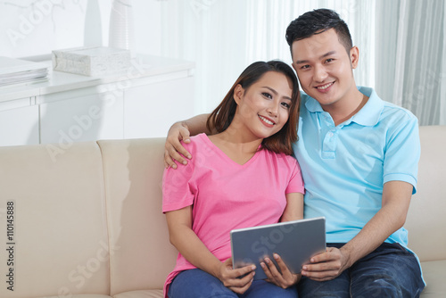 Happy Asian couple resting on sofa with digital tablet