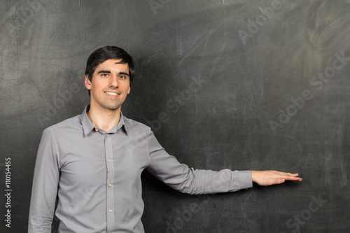 Portrait of young happy smiling teacher man standing near chalkboard background and showing something on it
