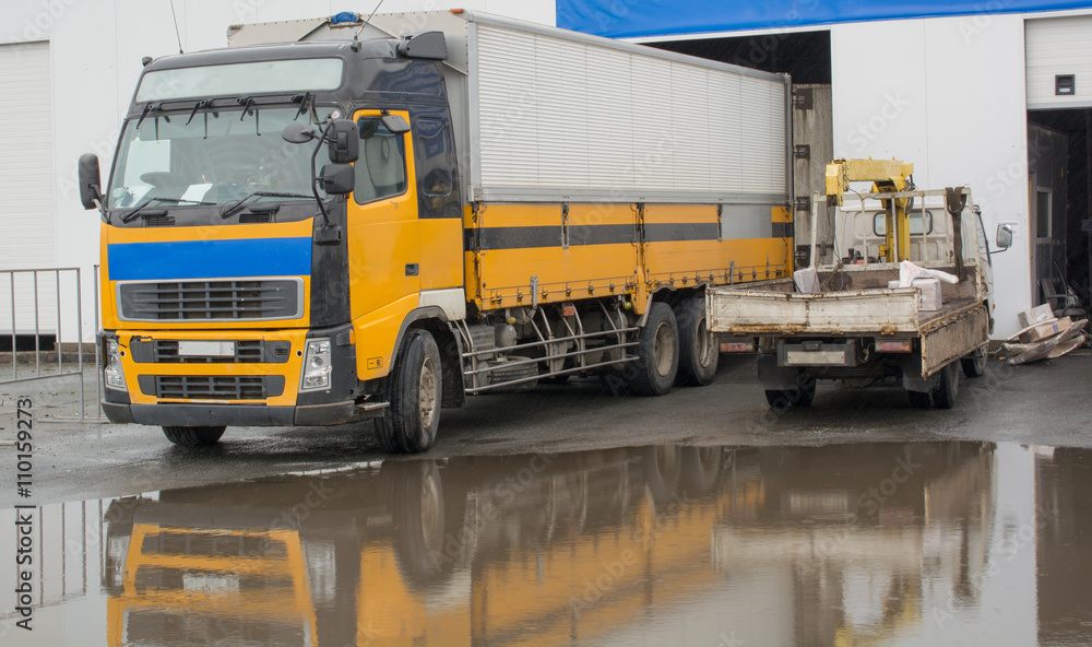The image of two trucks. One big yellow and one small standing near the warehouse.