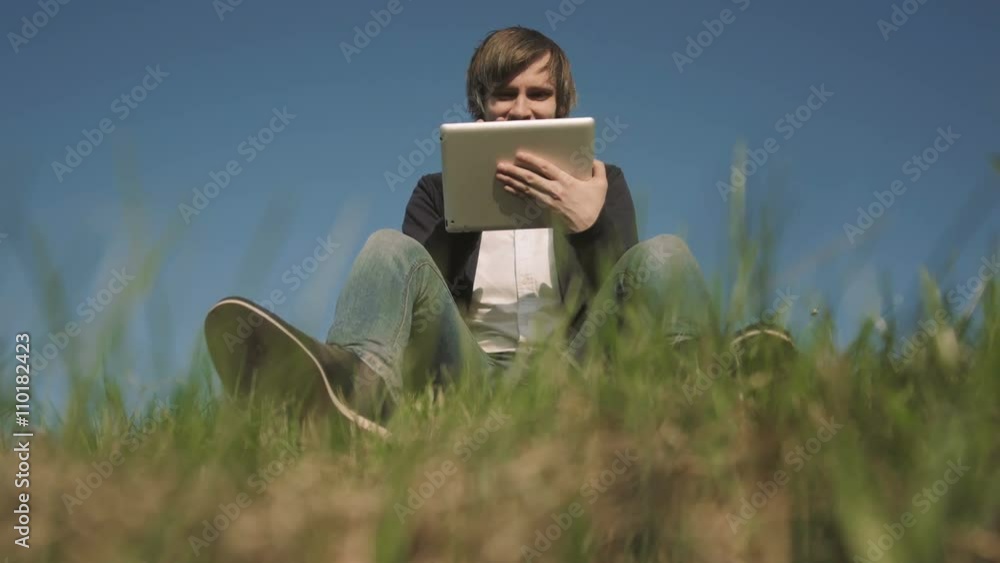 Bottom View of Handsome Hipster Using Tablet PC While Sitting On The Grass Bottom View of Handsome Hipster Using Tablet PC While Sitting On The Grass