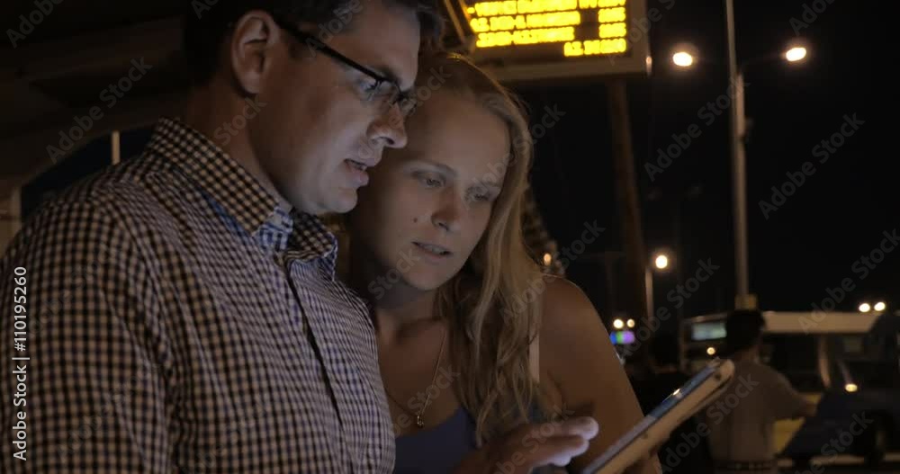 Young man and woman using touch pad on city bus station at night, information led display and people getting into the bus in background Young man and woman using touch pad on city bus station at night, information led display and people getting into the bus in background