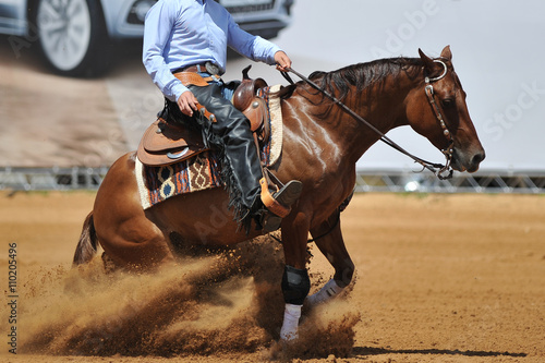 The side view of a rider in cowboy chaps and boots on a horseback running ahead in the dust.
