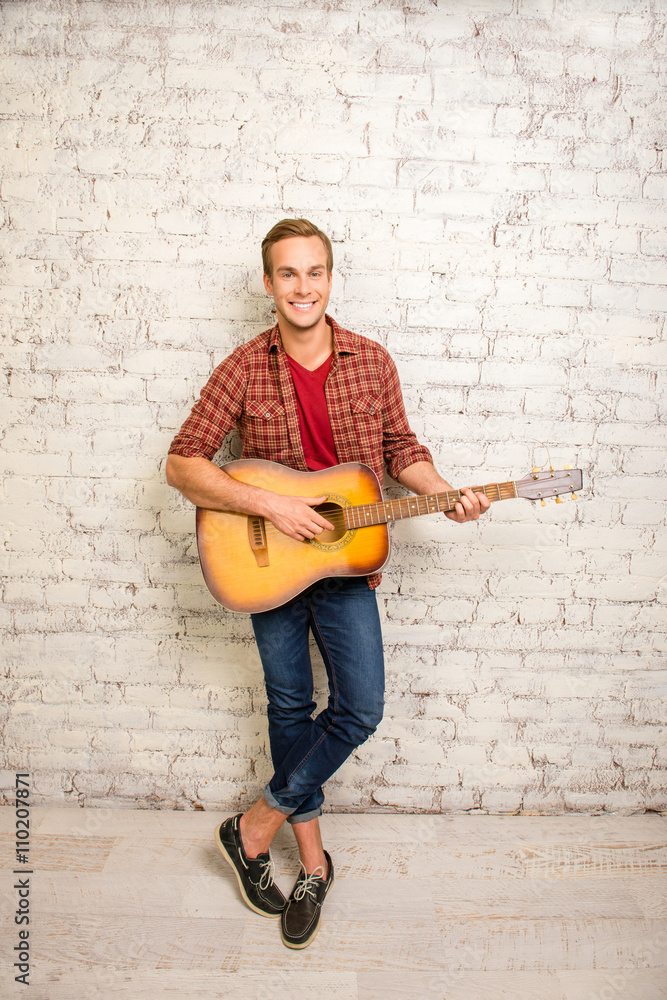 Smiling man standing near the wall and playing on the guitar