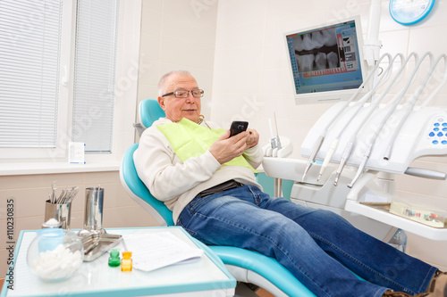 Old senior man sitting in a chair in dental clinic read or write sms on cell mobile phone.