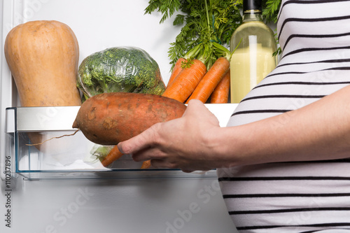 Pregnant woman opening fridge with vegetables and picking sweet potato