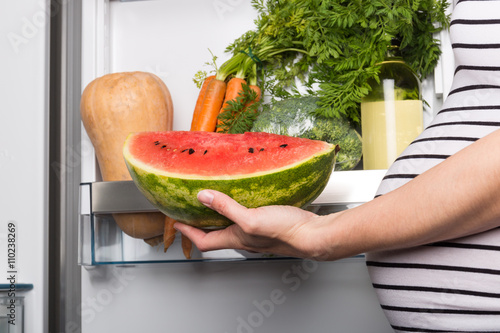 Pregnant woman opening fridge with vegetables and picking watermelon