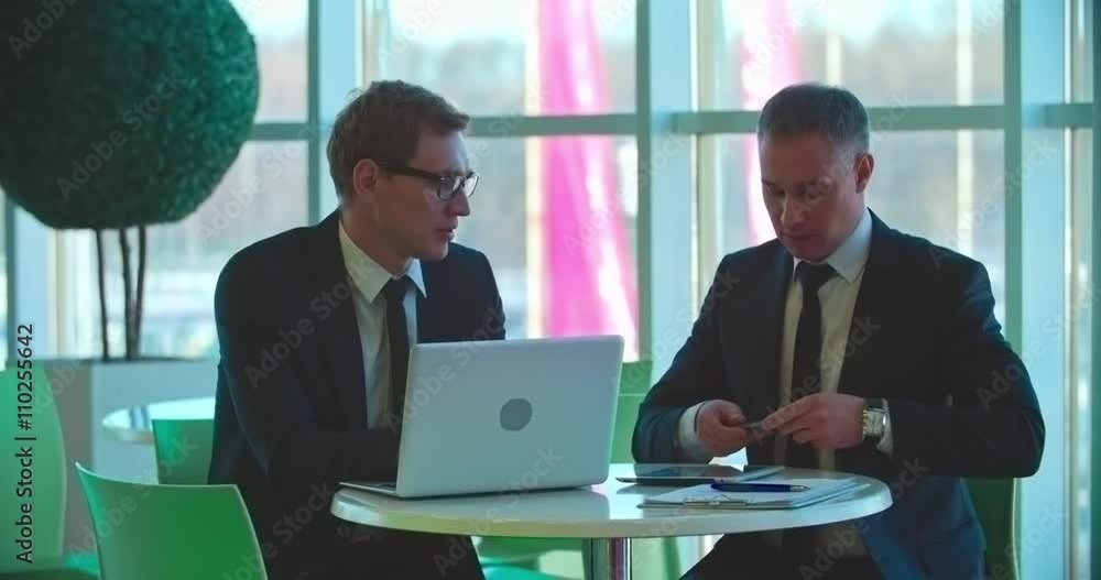 Two businessmen sitting at cafe table, using laptop, tablet and smartphone and talking Two businessmen sitting at cafe table, using laptop, tablet and smartphone and talking