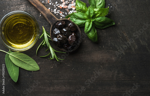 Greek black olives, fresh herbs and oil on dark rustic wooden background.