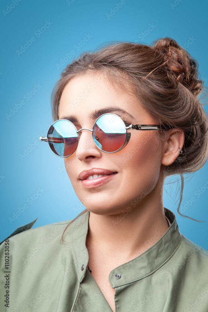 Closeup portrait of beautiful trendy hipster girl with hair buns smiling posing at camera wearing round sunglasses and green coat on blue background.Youth style,fashion. Closeup portrait of beautiful trendy hipster girl with hair buns smiling posing at camera wearing round sunglasses and green coat on blue background.Youth style,fashion.