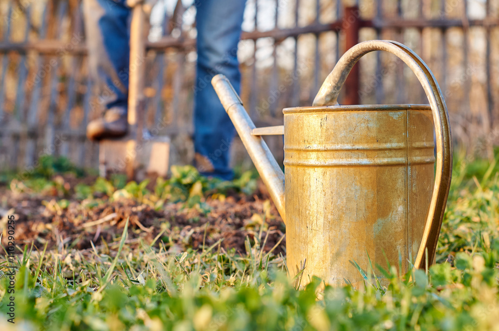 Metallic watering can in the foreground with a man digging garden beds using a spade in the background 