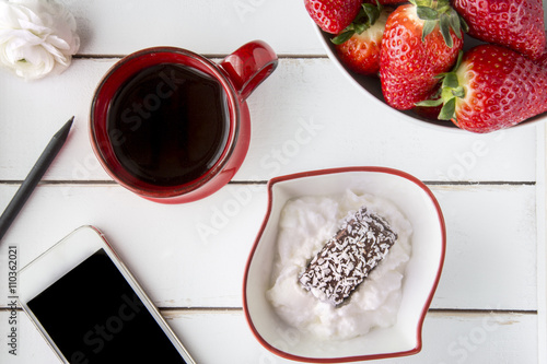 Overhead shot of a white wooden table, red coffee cup, white flower, smart phone, black pencil, strawberries, brown white biscuit on yoghurt
