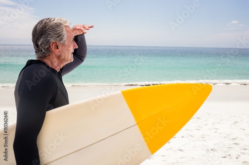 Senior man with surfboard shielding eyes at beach