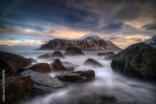 Rocks on coastline with Hustinden mountain and colorful clouds in background, Lofoten 