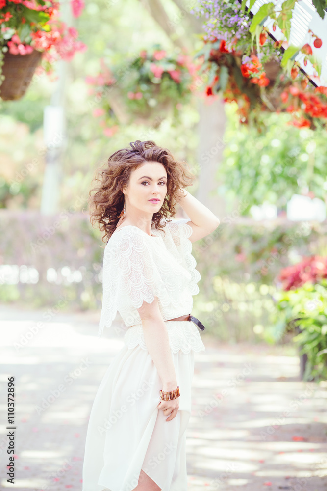 Portrait of beautiful smiling white Caucasian girl woman with long dark red brown hair, in white summer dress, standing in flowers in summer park garden, looking in camera Portrait of beautiful smiling white Caucasian girl woman with long dark red brown hair, in white summer dress, standing in flowers in summer park garden, looking in camera