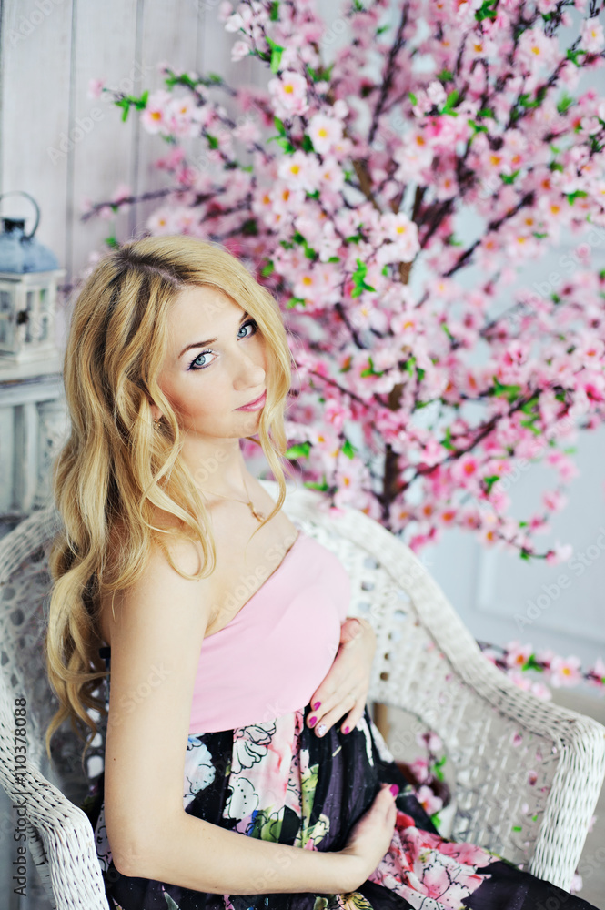 Young beautiful pregnant woman with long blond hair sitting in a wicker chair on the background of a decorative tree with pink flowers, look into the camera. Young beautiful pregnant woman with long blond hair sitting in a wicker chair on the background of a decorative tree with pink flowers, look into the camera.