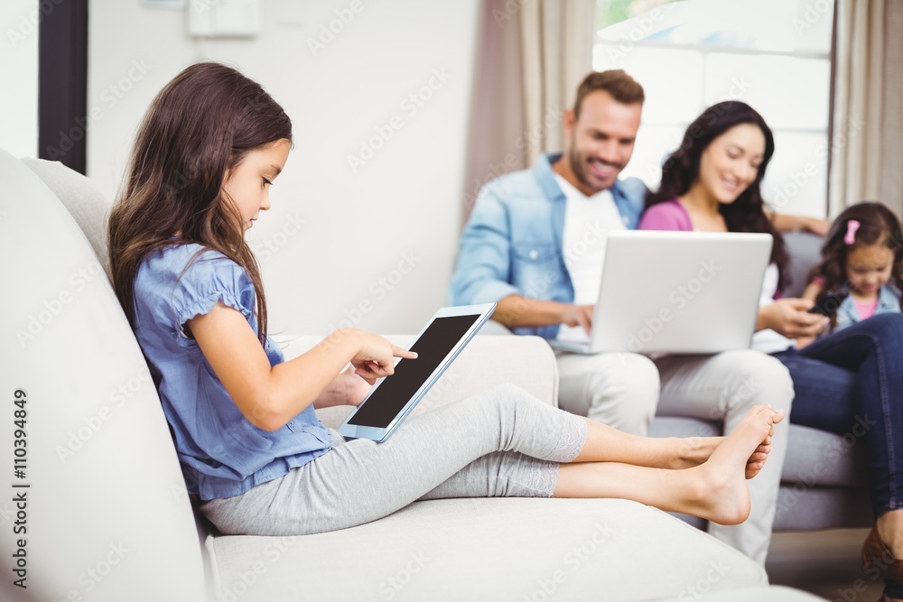 Girl using digital tablet on sofa while family in background Girl using digital tablet on sofa while family in background