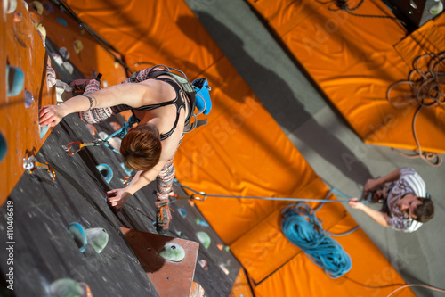 Strong young woman is climbing up with carbines and rope on an indoor rock-climbing wall. Man standing on the ground insuring the climber