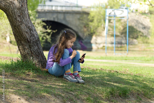 Cute little girl sitting on the grass in a park with a mobile phone in her hands and using modern smartphone