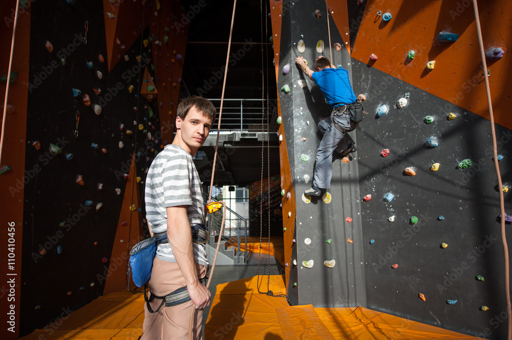 Man stands on the ground near rock wall indoors, looking at the camera and insuring the climber, using rope and belay device