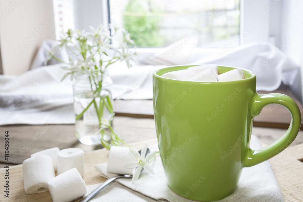 Hot cocoa chocolate with marshmallows in green cup on napkin and fresh spring white flowers on wooden table Hot cocoa chocolate with marshmallows in green cup on napkin and fresh spring white flowers on wooden table