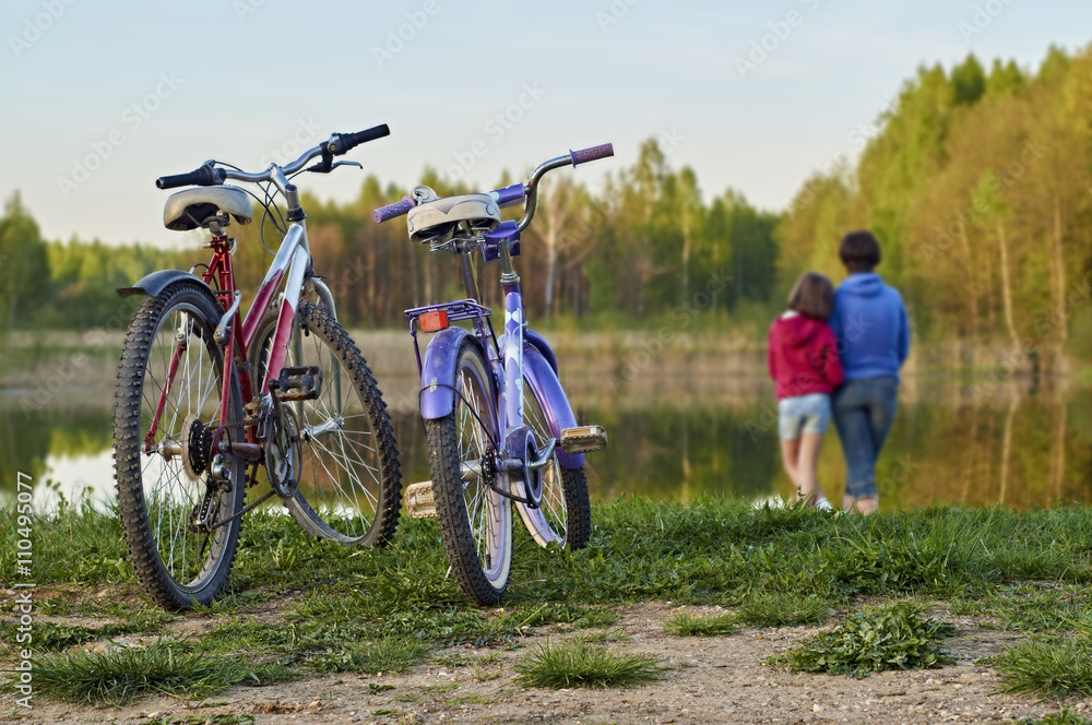 Two bicycles in the foreground and mother with daughter standing at the lake shore in the blurred background Two bicycles in the foreground and mother with daughter standing at the lake shore in the blurred background
