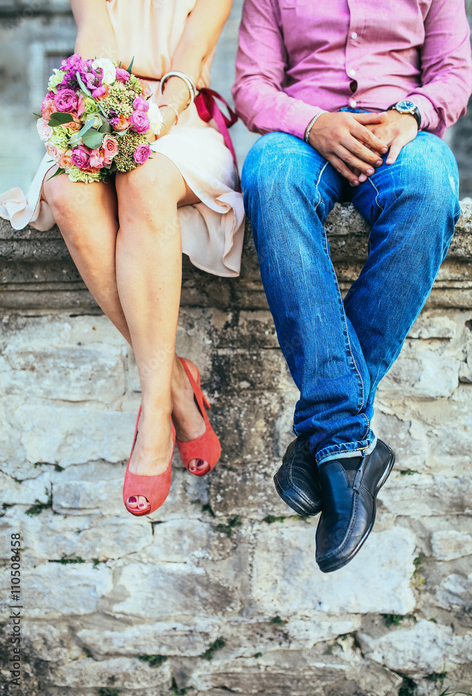 A couple sitting on a stone wall in jeans wearing shoes. Shot from the legs down.