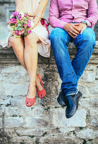 A couple sitting on a stone wall in jeans wearing shoes. Shot from the legs down.