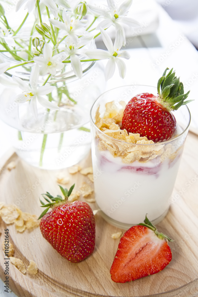 Homemade healthy dessert in a glass with yogurt, fresh strawberry and corn flakes for breakfast with fresh white flowers on wooden table, closeup Homemade healthy dessert in a glass with yogurt, fresh strawberry and corn flakes for breakfast with fresh white flowers on wooden table, closeup