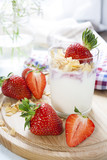 Homemade healthy dessert in a glass with yogurt, fresh strawberry and corn flakes for breakfast with fresh white flowers and checkered napkin on wooden table, closeup