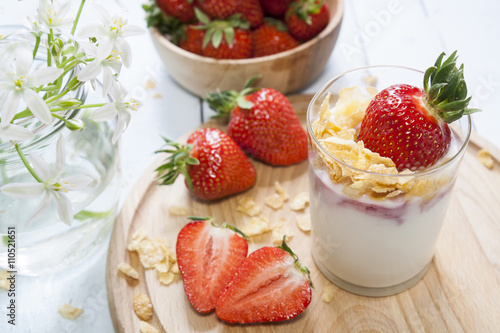Homemade healthy dessert in a glass with yogurt  fresh strawberry and corn flakes for breakfast with fresh white flowers on wooden table  closeup