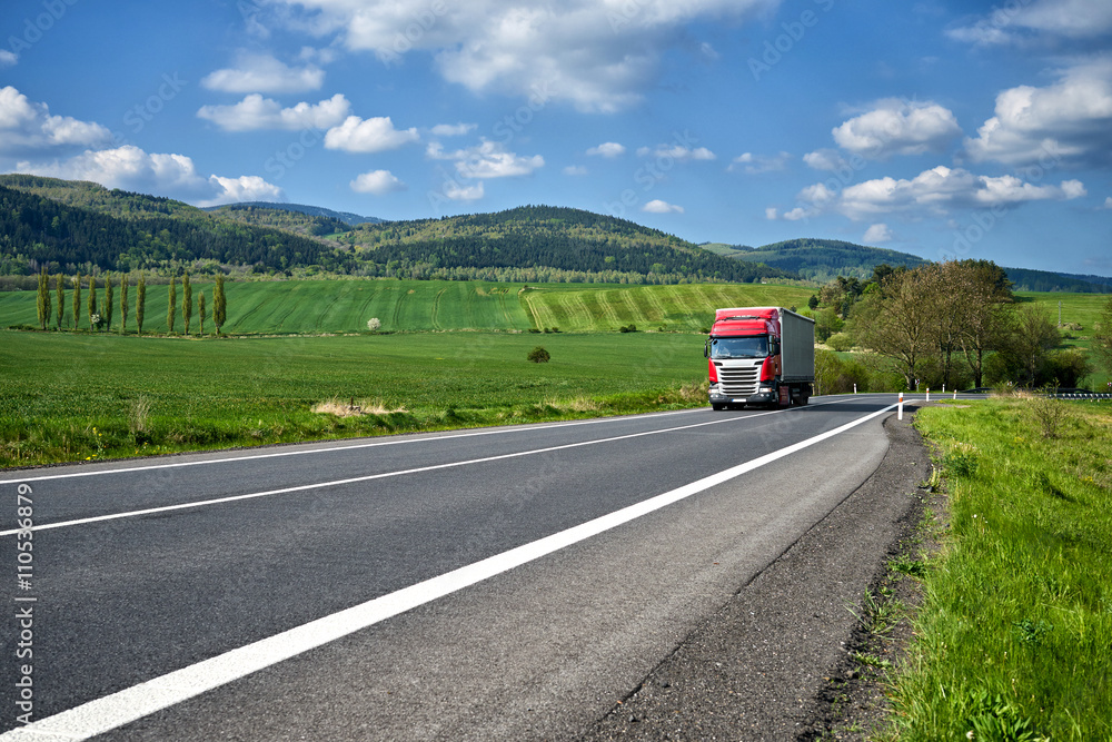 Rural landscape with asphalt road and oncoming red truck. Green fields and wooded mountains in the background. Sunny spring day with blue skies and white clouds.