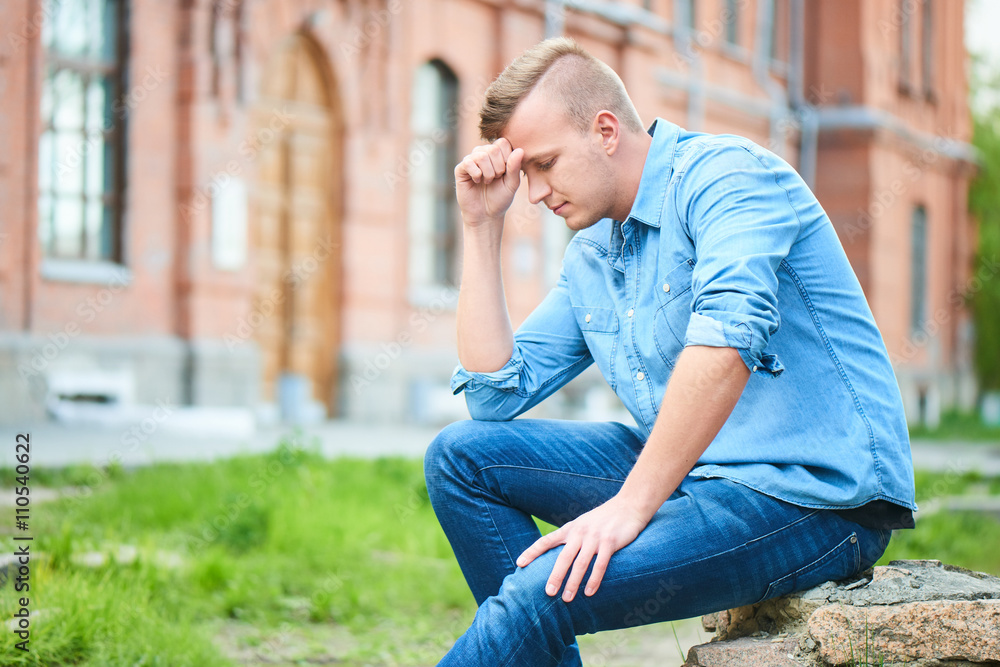 Portrait of a smiling student sitting in a denim shirt and holding a head in his hand, against the background of the school building of brick Portrait of a smiling student sitting in a denim shirt and holding a head in his hand, against the background of the school building of brick